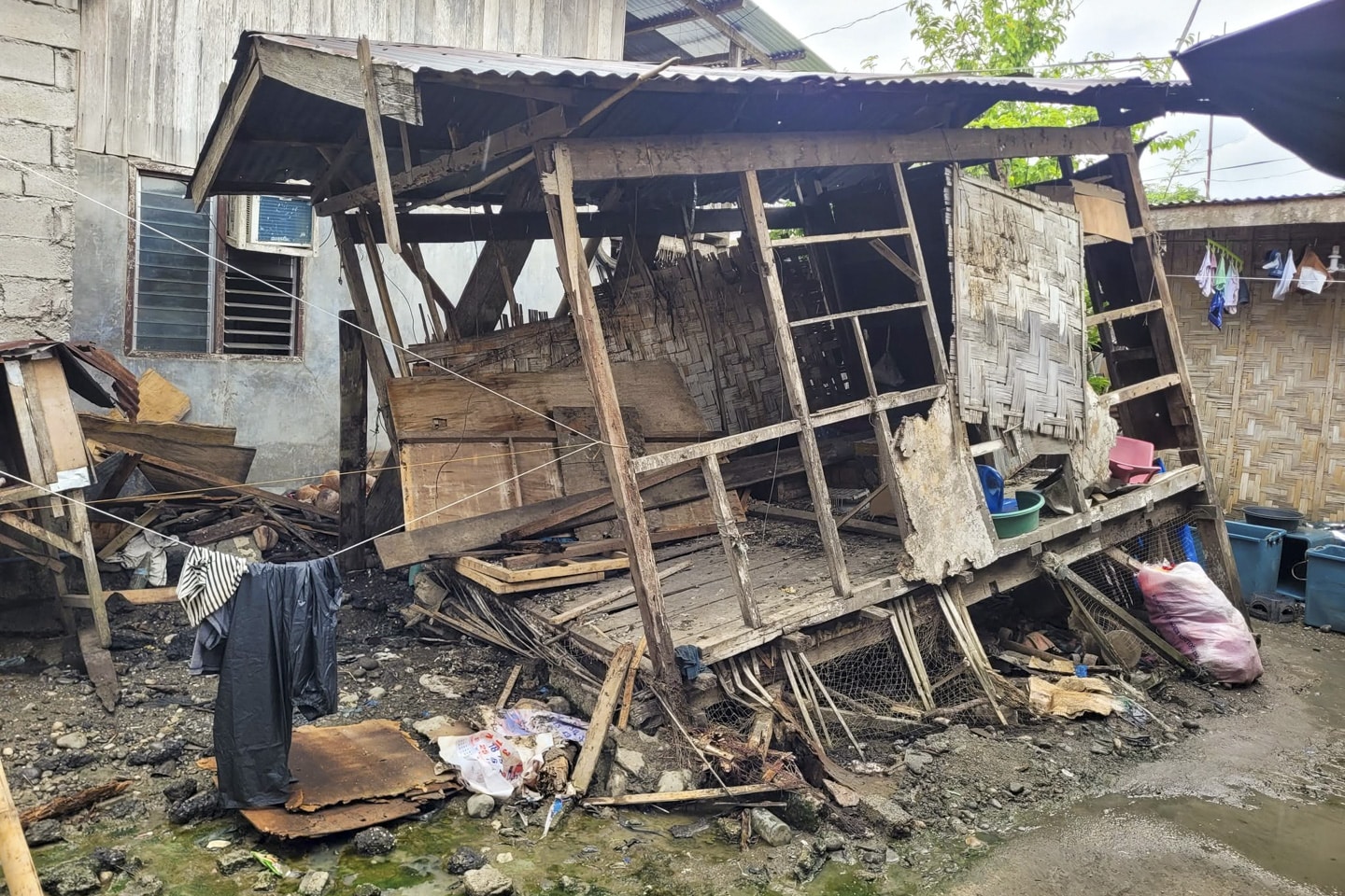 "A small, fragile house made of basic materials like wood and corrugated metal sheets, heavily damaged and partially collapsed from the earthquake. The structure is leaning precariously, surrounded by debris, highlighting the vulnerability of families displaced by the disaster."